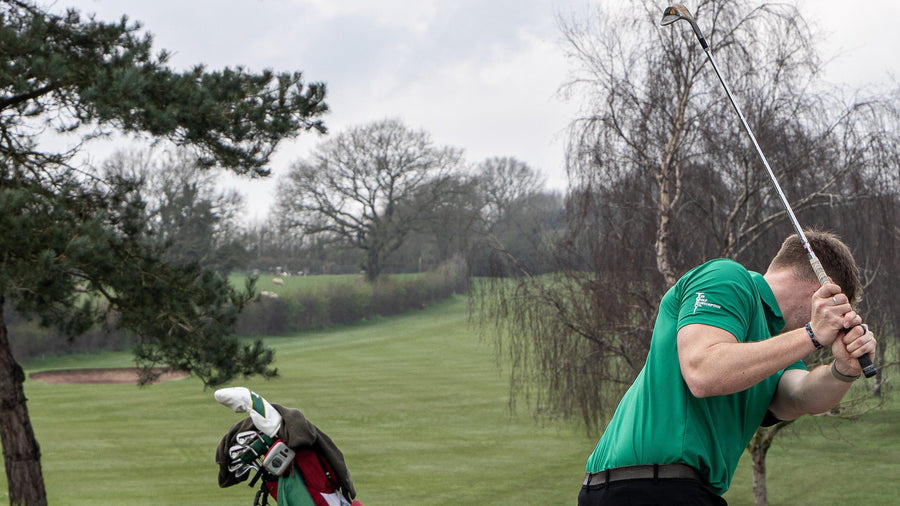 Golfer in green shirt swinging a club on a golf course with trees and grass in the background.